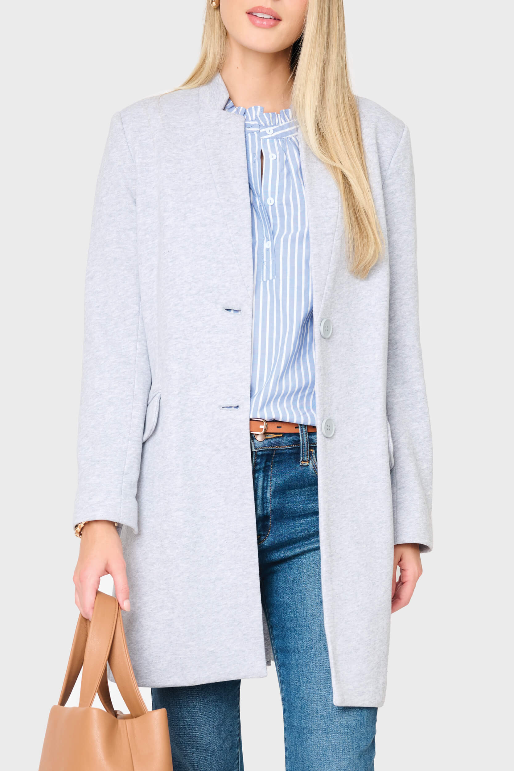 Woman wearing a light gray coat over a striped shirt with blue jeans, holding a brown handbag on a plain background.