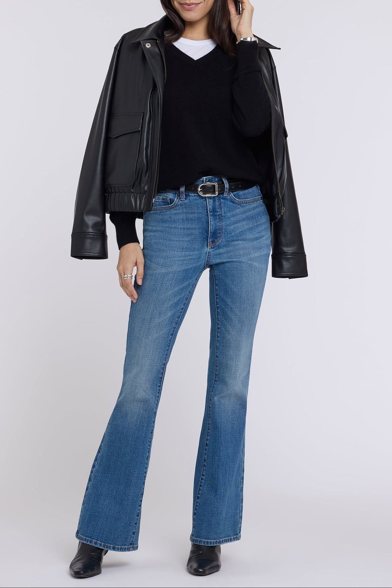 A woman modeling a black faux-leather moto jacket, black sweater, blue flare jeans, and black boots against a light gray studio background.