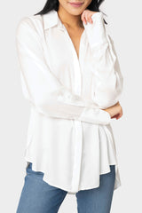 A woman wearing a white button-front blouse with long sleeves, a point collar, and button cuffs. The blouse is styled in a satin effect fabric and is shown in an open, casual look.