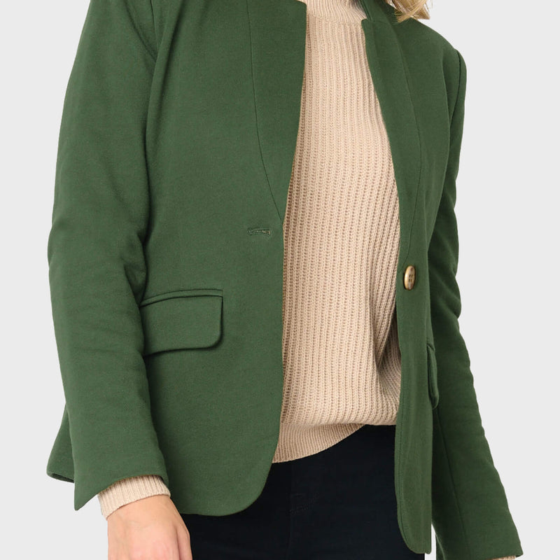 A woman wearing a dark green tailored blazer over a beige ribbed turtleneck sweater and black pants, shown from the mouth down against a light gray studio background.