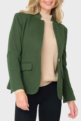 A woman wearing a dark green tailored blazer over a beige ribbed turtleneck sweater and black pants, shown from the mouth down against a light gray studio background.