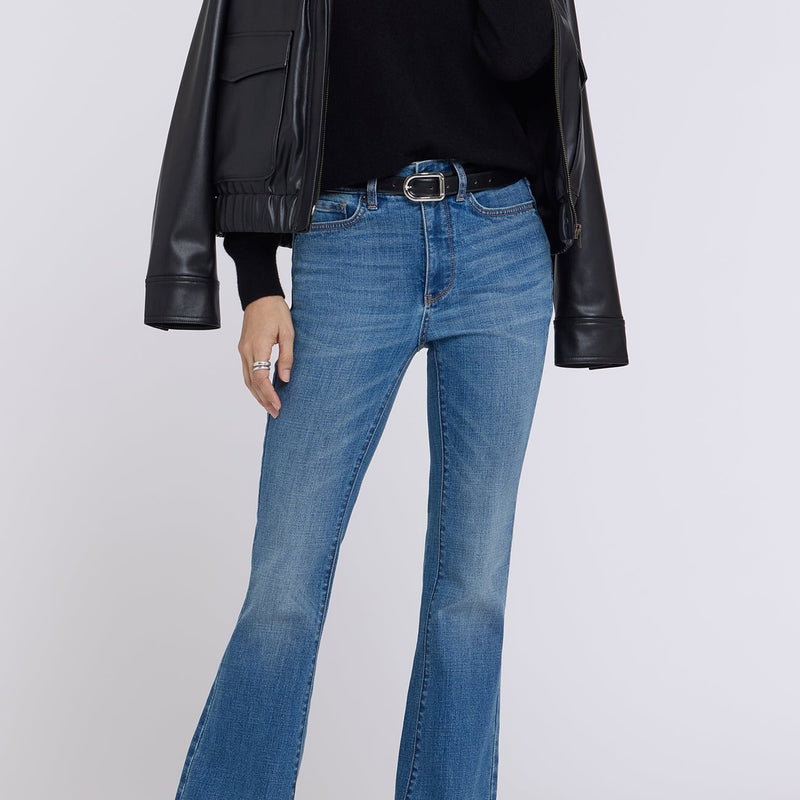 A woman modeling a black faux-leather moto jacket, black sweater, blue flare jeans, and black boots against a light gray studio background.