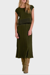 A woman modeling a matching olive green two-piece knit set consisting of a short-sleeve top with a wide waistband and a pleated midi skirt, paired with tan suede boots, against a textured beige studio backdrop.
