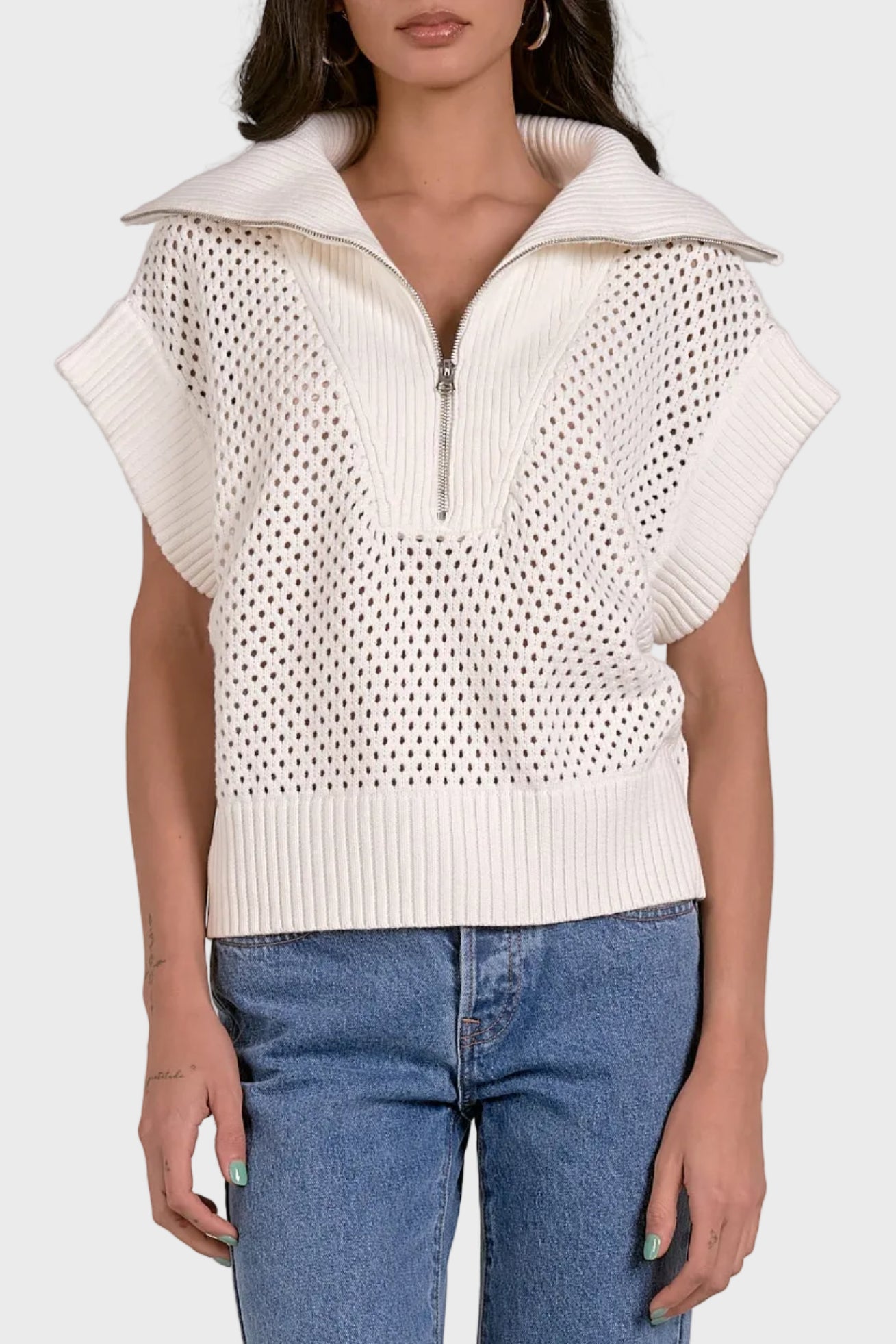 Woman wearing a white sleeveless knit top with a zipper and blue jeans on a light gray background