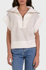 Woman wearing a white sleeveless knit top with a zipper and blue jeans on a light gray background