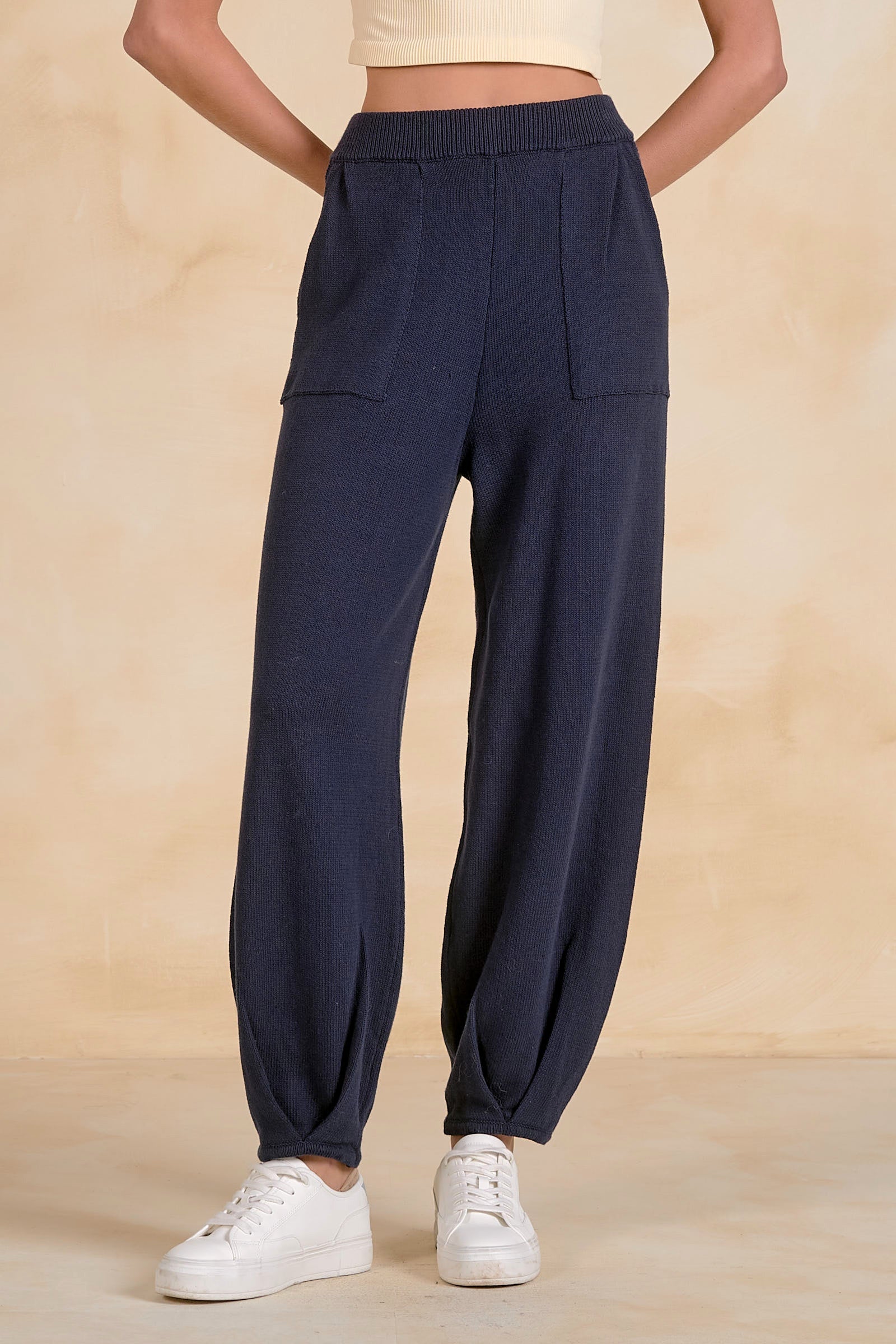 A person wearing dark navy blue lounge pants with a wide elastic waistband, side pockets, and gathered cuffs, paired with white sneakers against a warm beige studio backdrop.