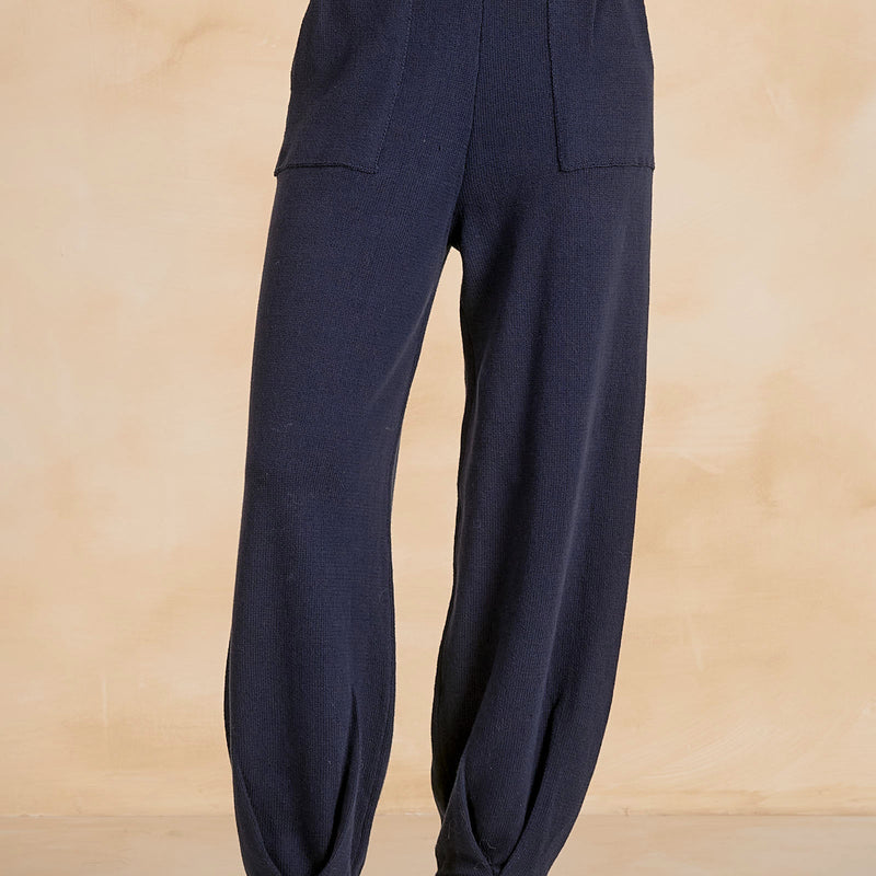 A person wearing dark navy blue lounge pants with a wide elastic waistband, side pockets, and gathered cuffs, paired with white sneakers against a warm beige studio backdrop.