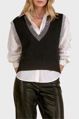 A woman modeling a black ribbed knit sweater vest with gray striped trim over a white button-down shirt and black leather pants, set against a warm beige textured background.