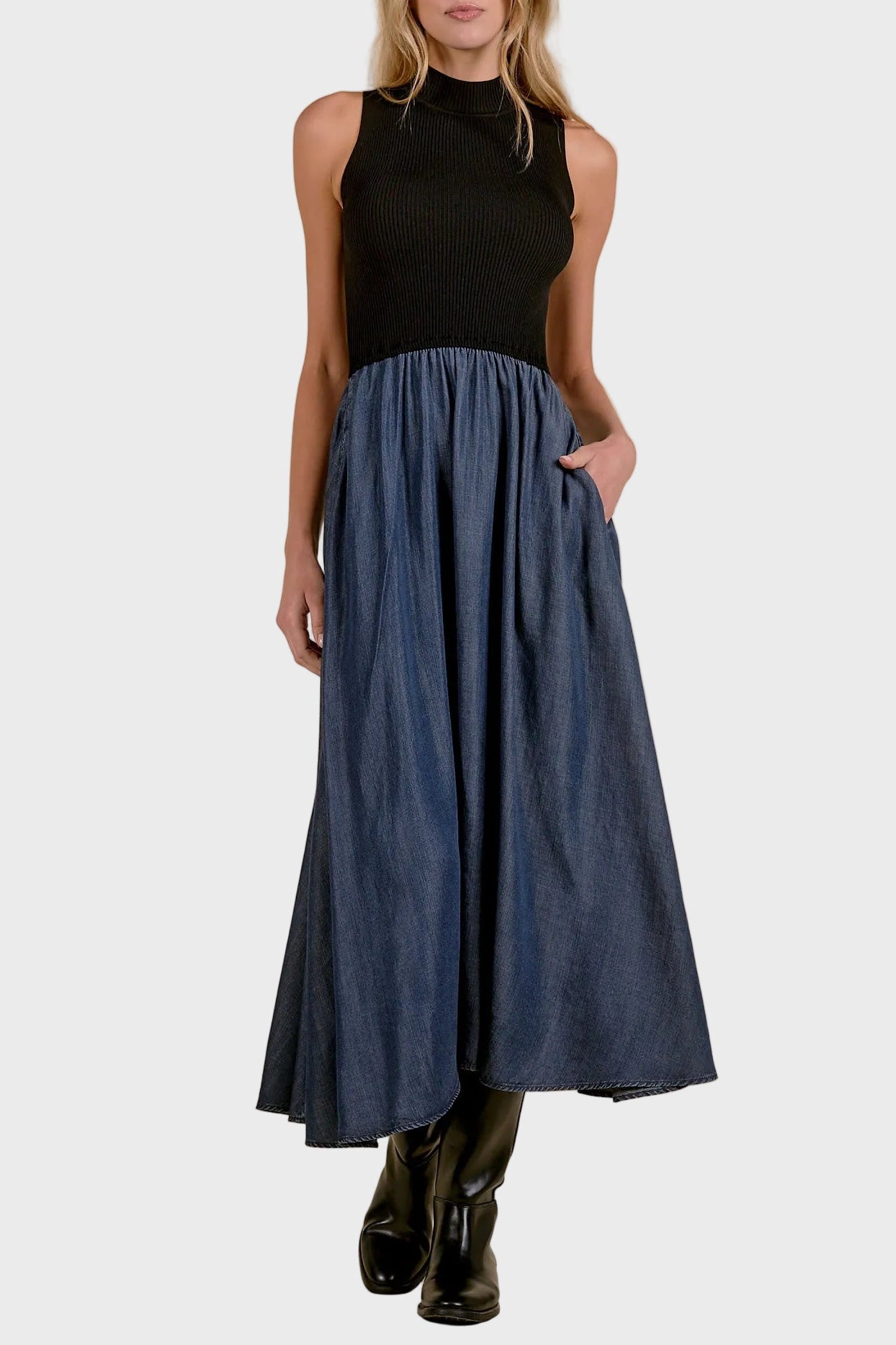 A woman modeling a sleeveless black mock-neck top paired with a high-waisted, flowing blue denim skirt with side pockets, and black boots, set against a textured beige studio backdrop.
